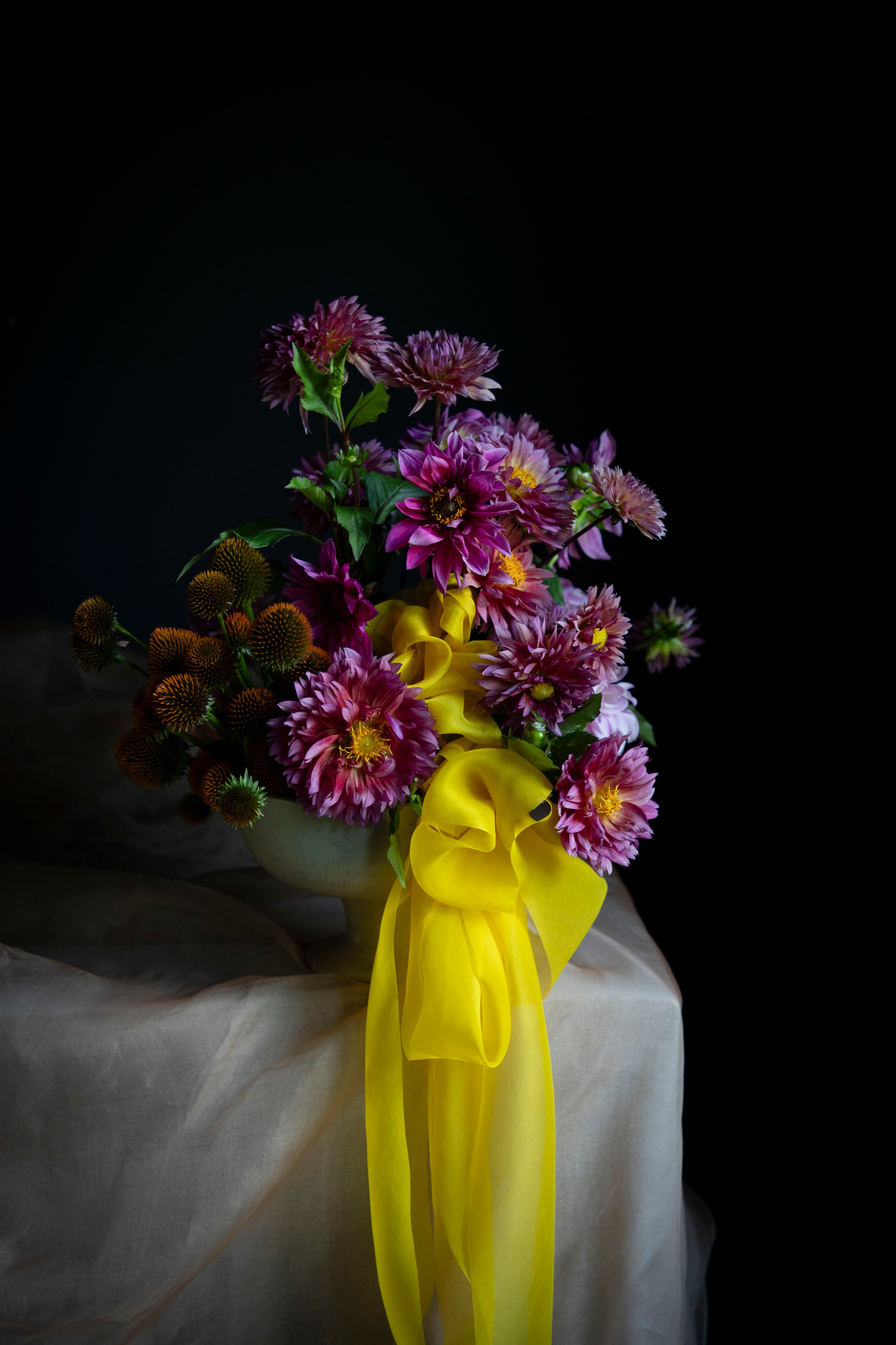 A bouquet of purple and pink dahlia flowers with green stems, tied with a bright yellow ribbon, set against a dark black background with draped white fabric below.