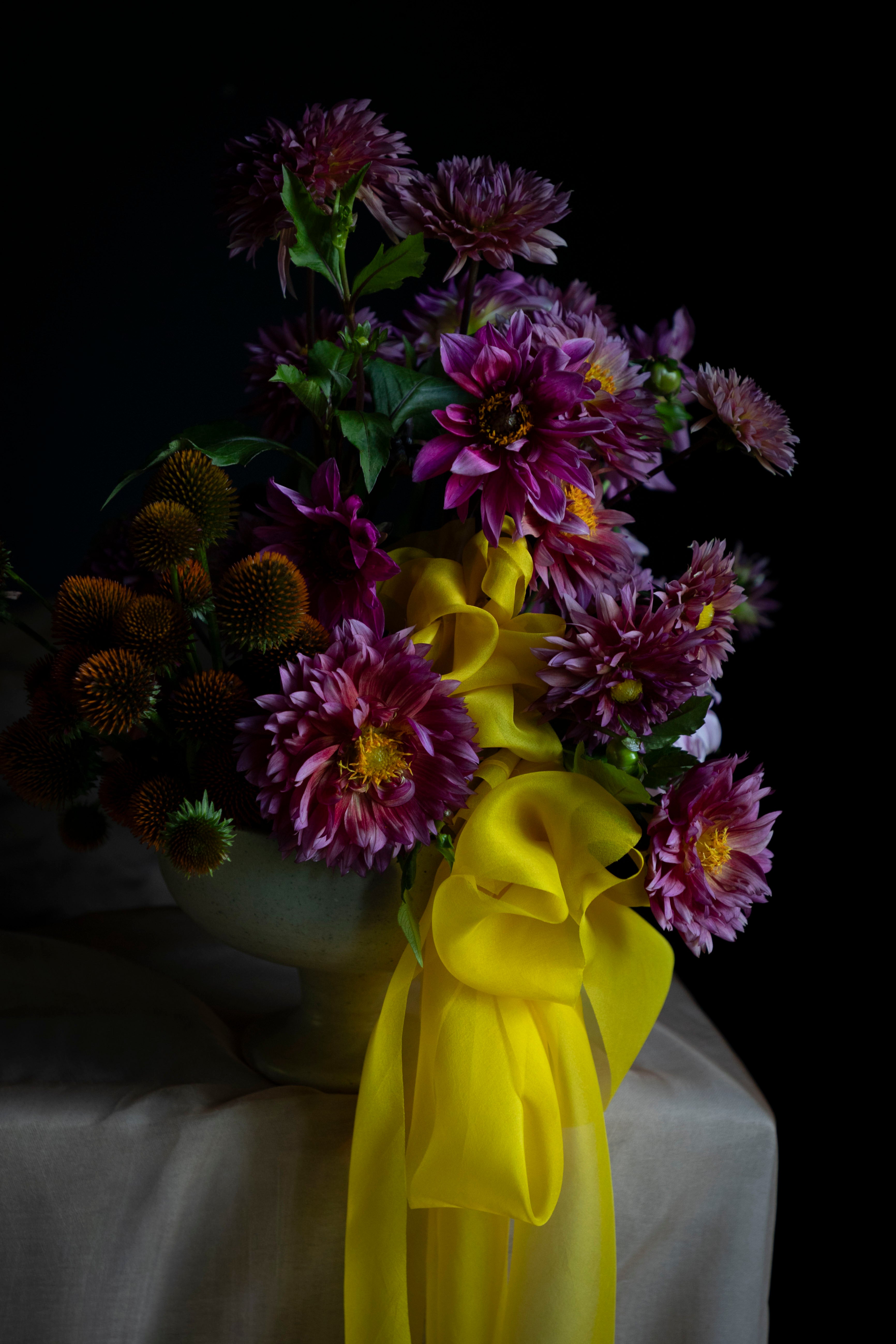 A floral arrangement of purple chrysanthemums and orange coneflowers in a pale green ceramic vase, tied with a large bright yellow ribbon, set against a dark background on a draped white cloth.