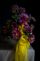 A floral arrangement of purple chrysanthemums and orange coneflowers in a pale green ceramic vase, tied with a large bright yellow ribbon, set against a dark background on a draped white cloth.
