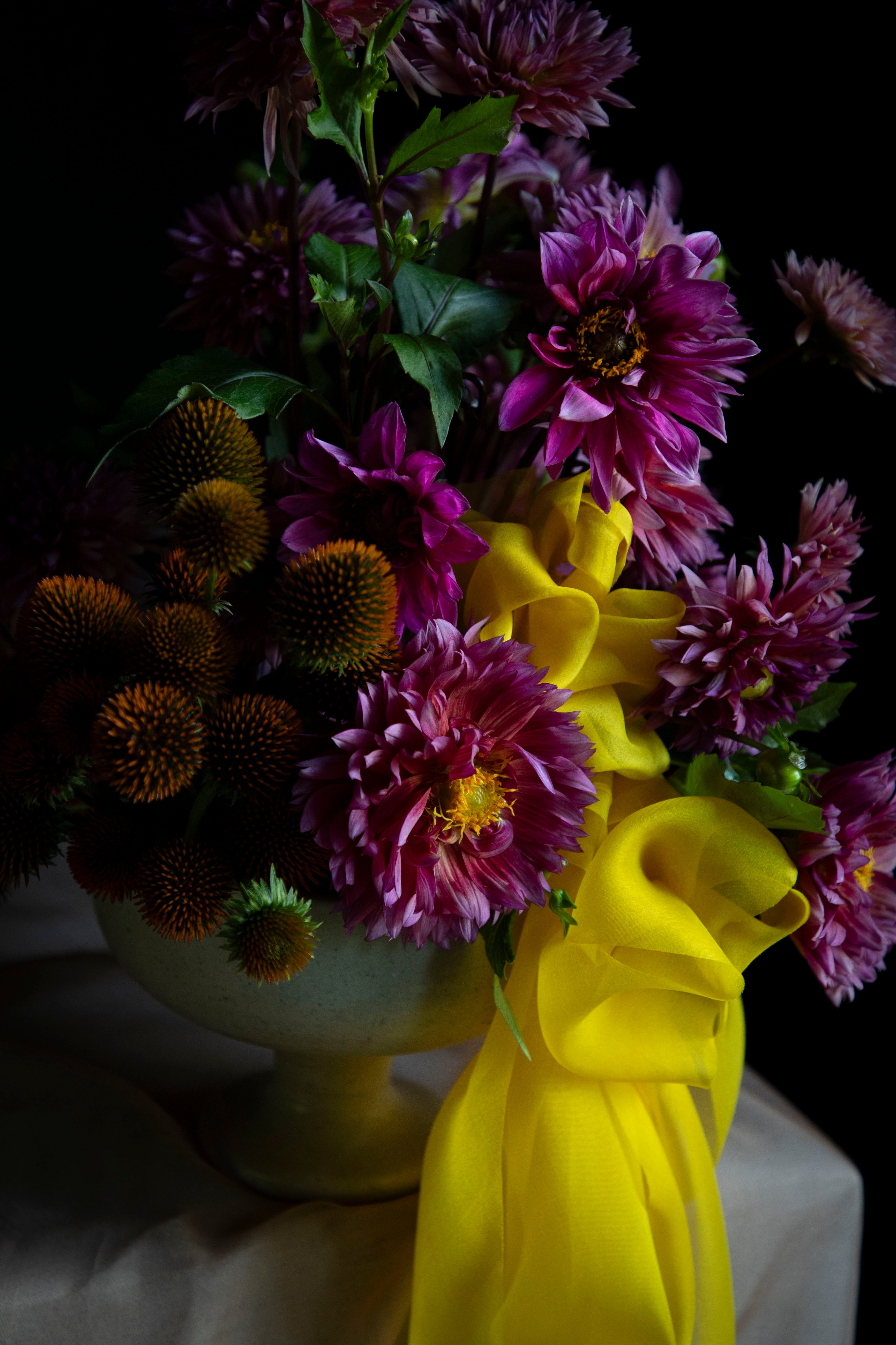 A vibrant floral arrangement featuring purple dahlia flowers, orange coneflowers, and a bright yellow satin ribbon, presented in a pale yellow vase against a dark background.