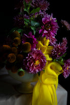 A vibrant floral arrangement featuring purple dahlia flowers, orange coneflowers, and a bright yellow satin ribbon, presented in a pale yellow vase against a dark background.