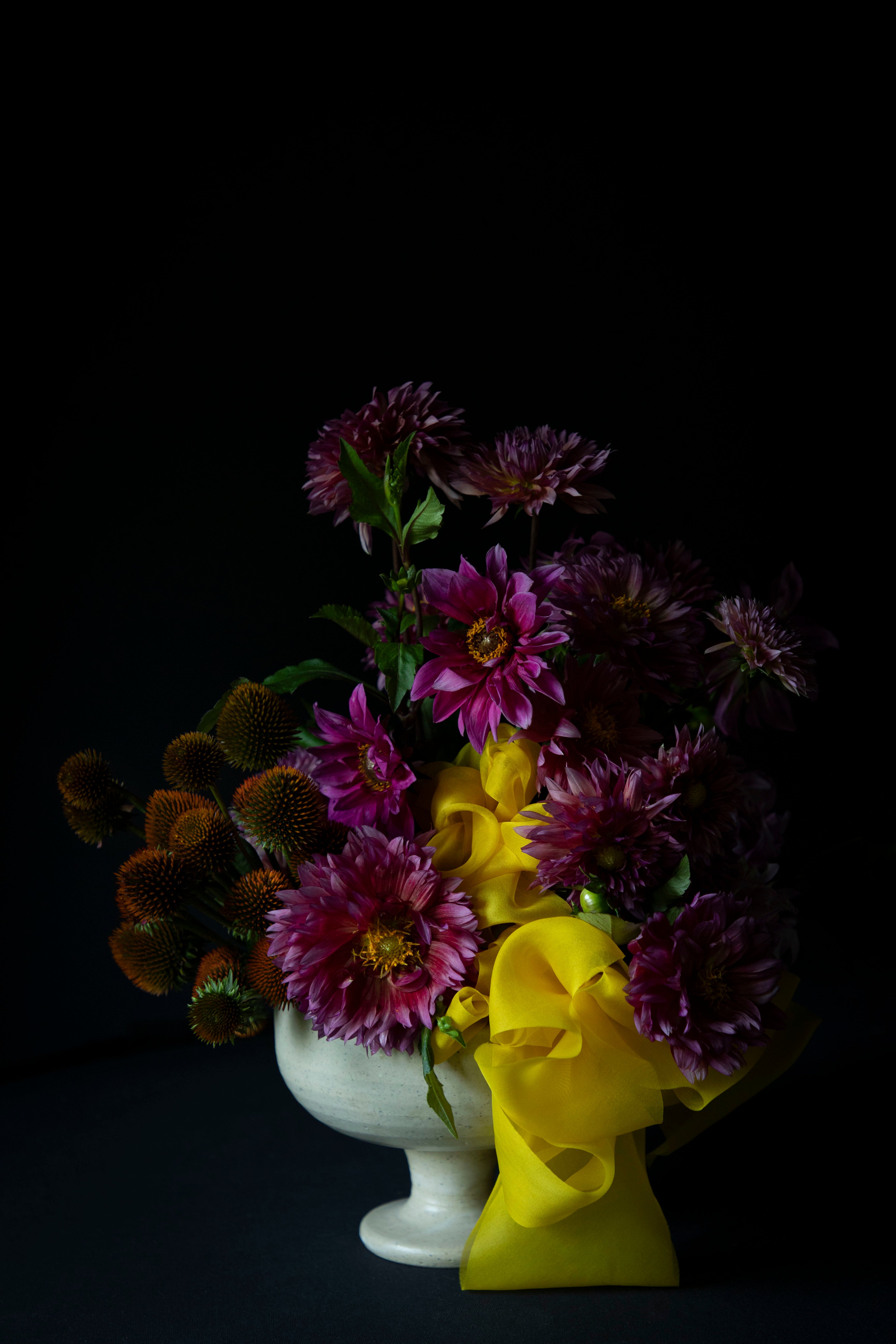 A floral arrangement of purple dahlia flowers, dark orange thistle-like blooms, and a bright yellow ribbon bow, presented in a white pedestal vase against a solid black background.