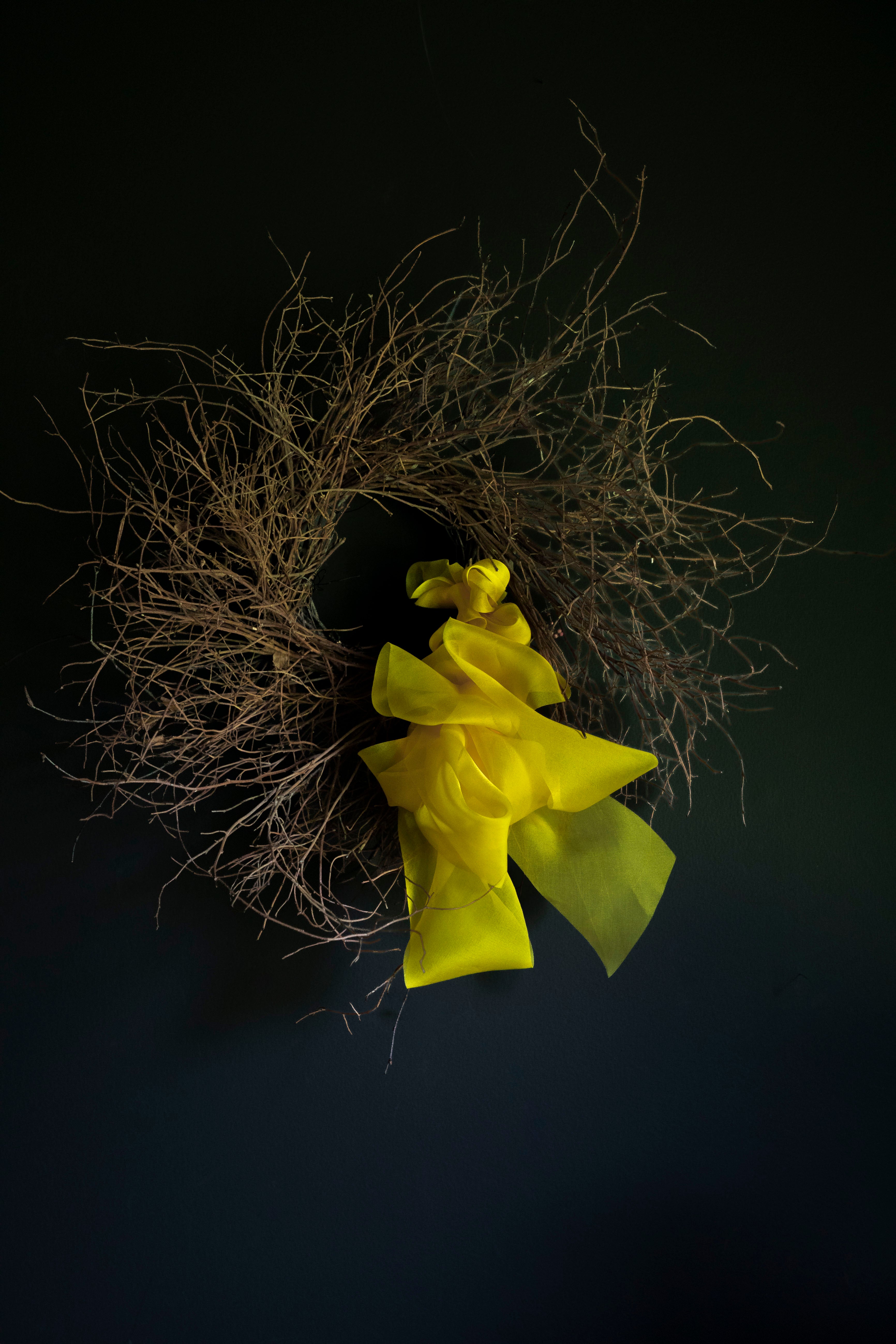 A rustic decorative wreath made of dried brown twigs and branches, accented with a large bright yellow fabric bow, set against a dark black background.