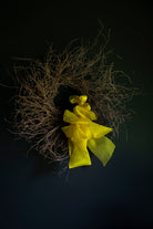 A rustic decorative wreath made of dried brown twigs and branches, accented with a large bright yellow fabric bow, set against a dark black background.
