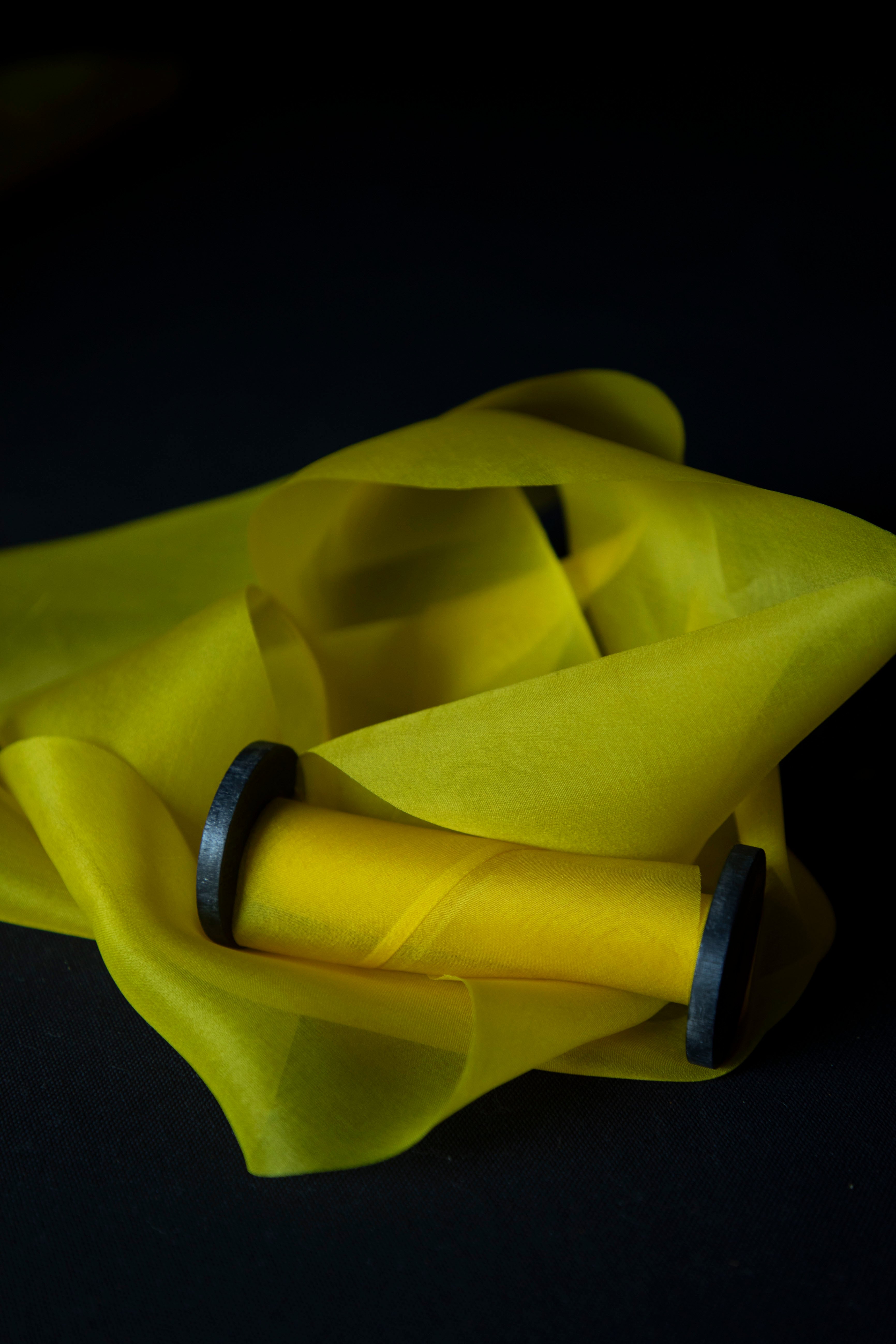 A bright yellow sheer fabric ribbon draped over a wooden spool, set against a dark black background.