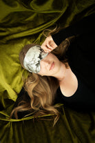woman laying of a bed with green velvet sheets wearing a marbled pattern silk sleep eye mask by silk & Willow