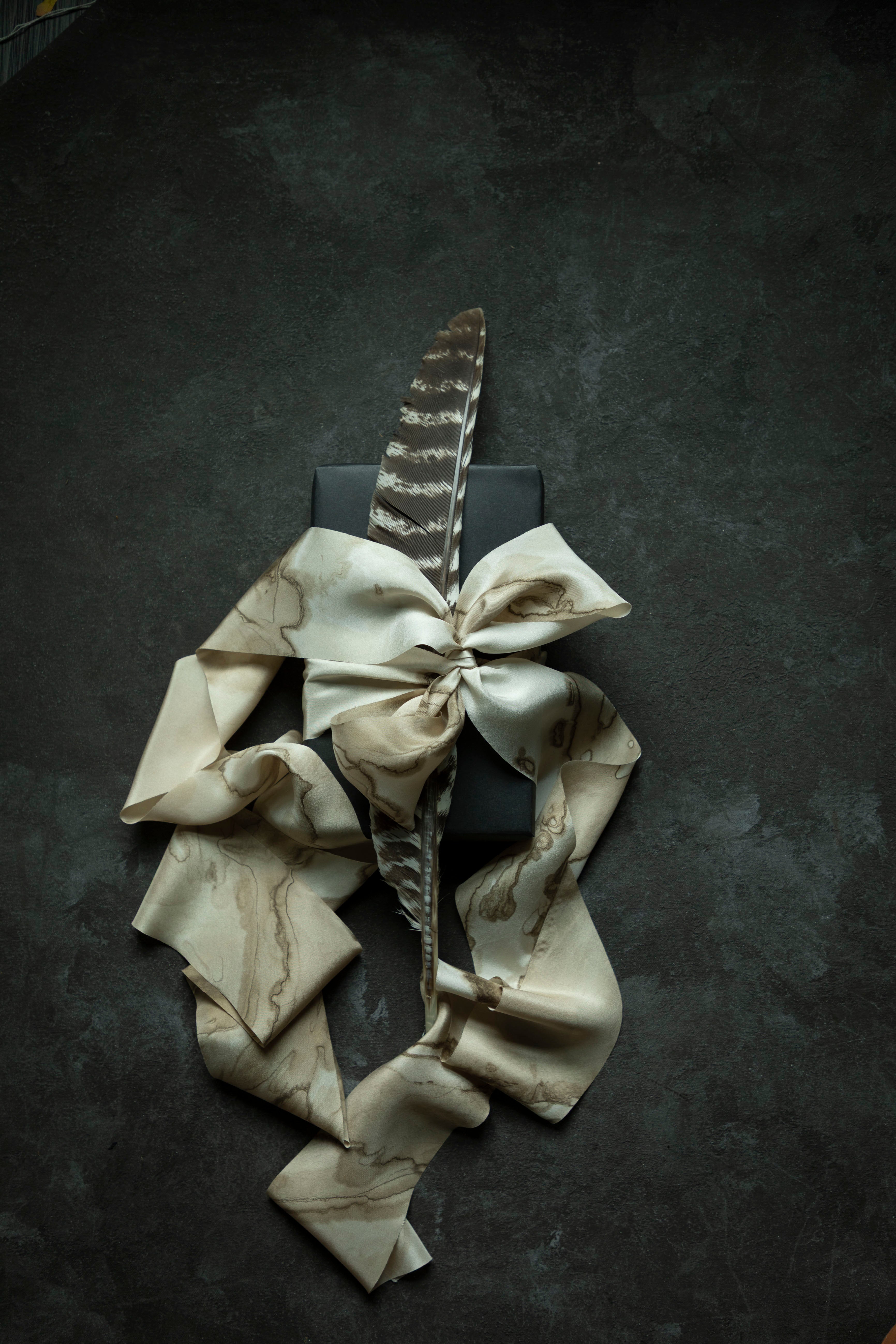 A cream and beige silk ribbon with a woodgrain marbled pattern, styled with a large bow and a single brown feather on top of a black rectangular box, set against a dark textured background.