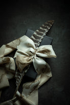 A styled flat-lay of a tan silk ribbon with brown abstract veining, like woodgrain, tied into a large bow around a dark gray gift box, with a brown and black patterned feather placed on top, set against a dark textured background.