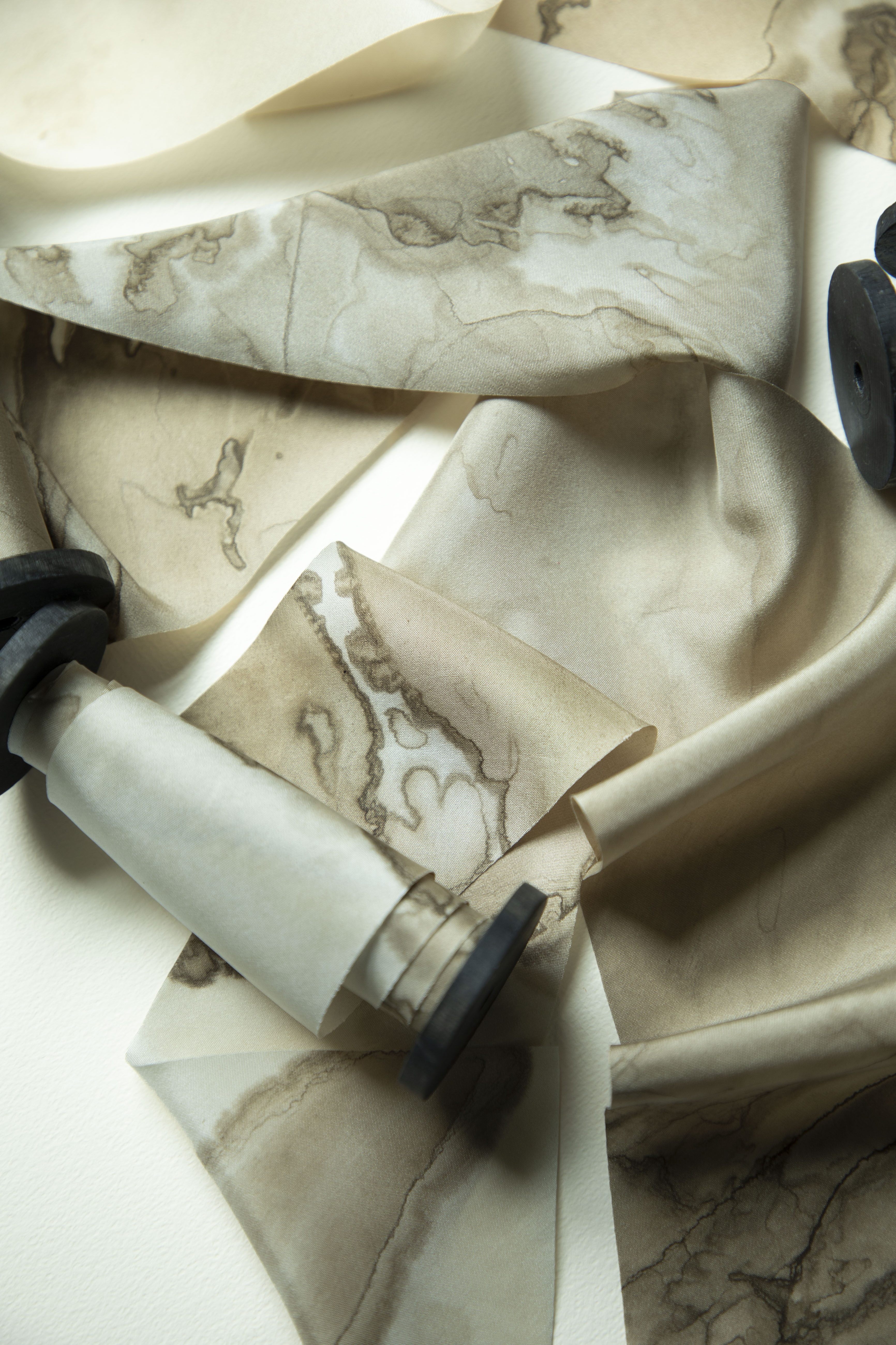 A close-up of draped silk ribbon with a beige and cream marble or stone-like pattern, arranged on a light surface with a few dark round props partially visible.