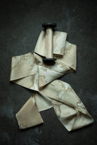 A beige silk ribbon with a brown marble pattern, draped artistically around a black spool against a dark textured background.