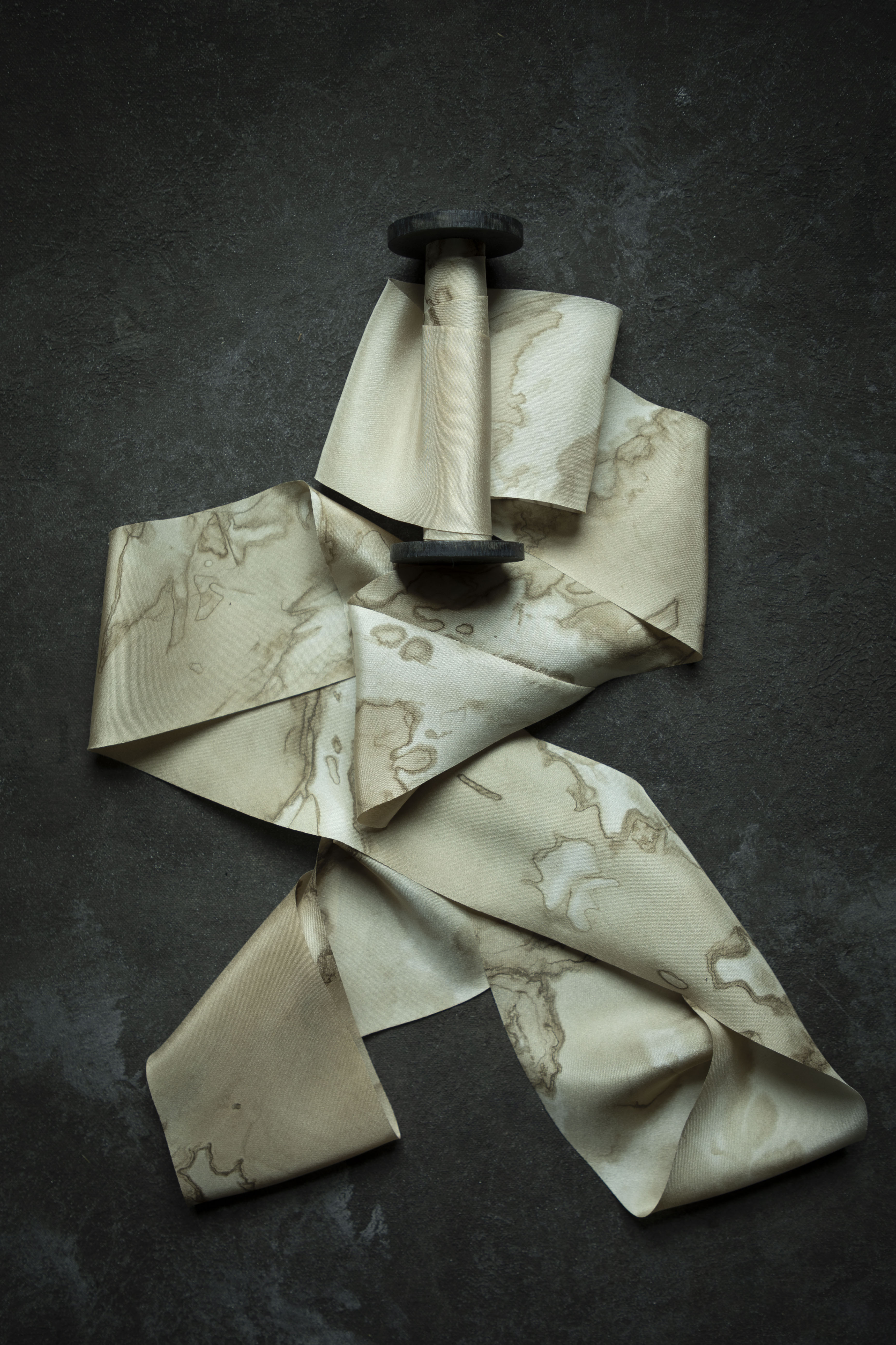 A beige silk ribbon with a brown marble pattern, draped artistically around a black spool against a dark textured background.