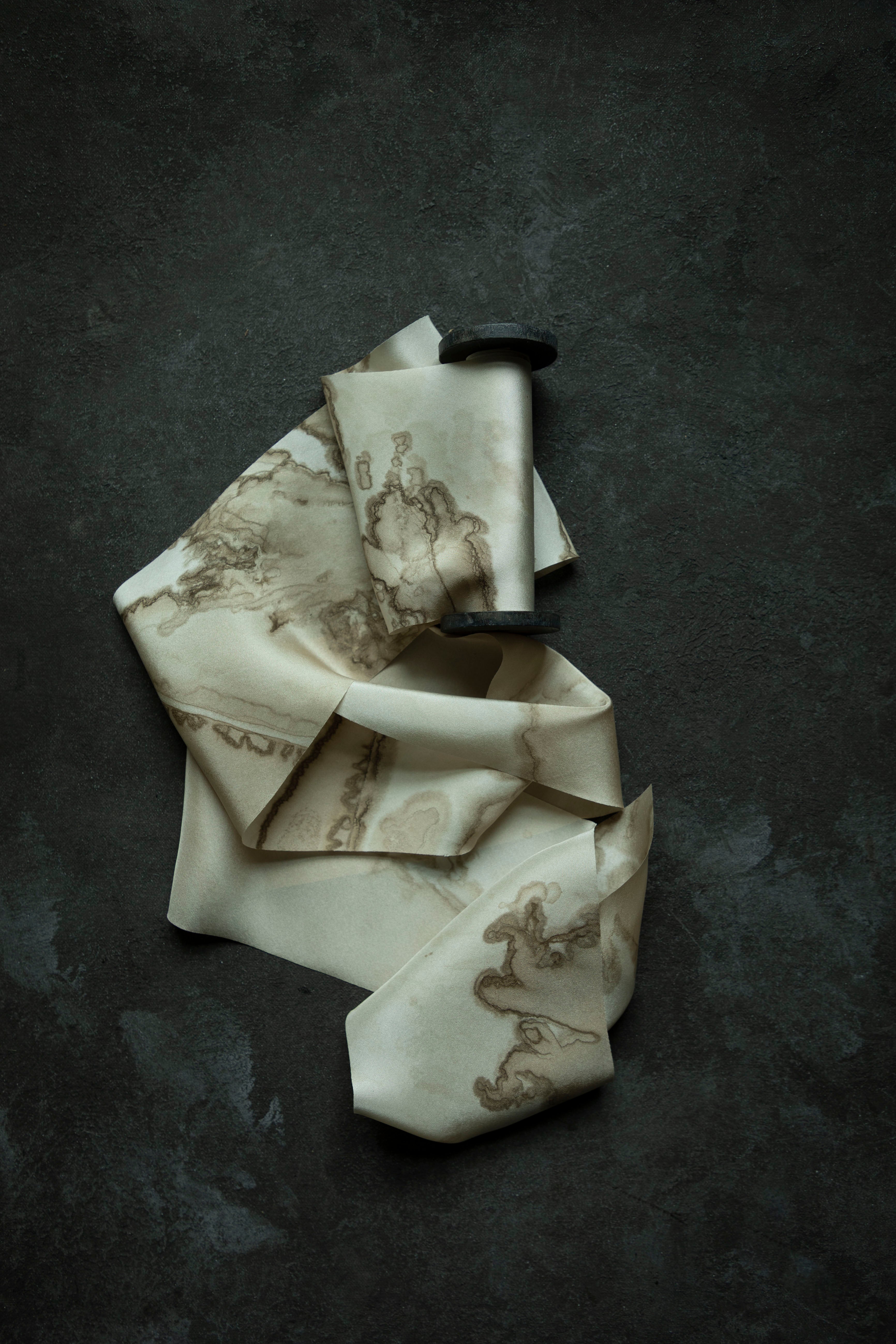 A cream-colored silk scarf with a brown abstract ink-blot pattern, draped artistically on a dark textured slate surface.