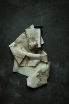 A cream-colored silk scarf with a brown abstract ink-blot pattern, draped artistically on a dark textured slate surface.