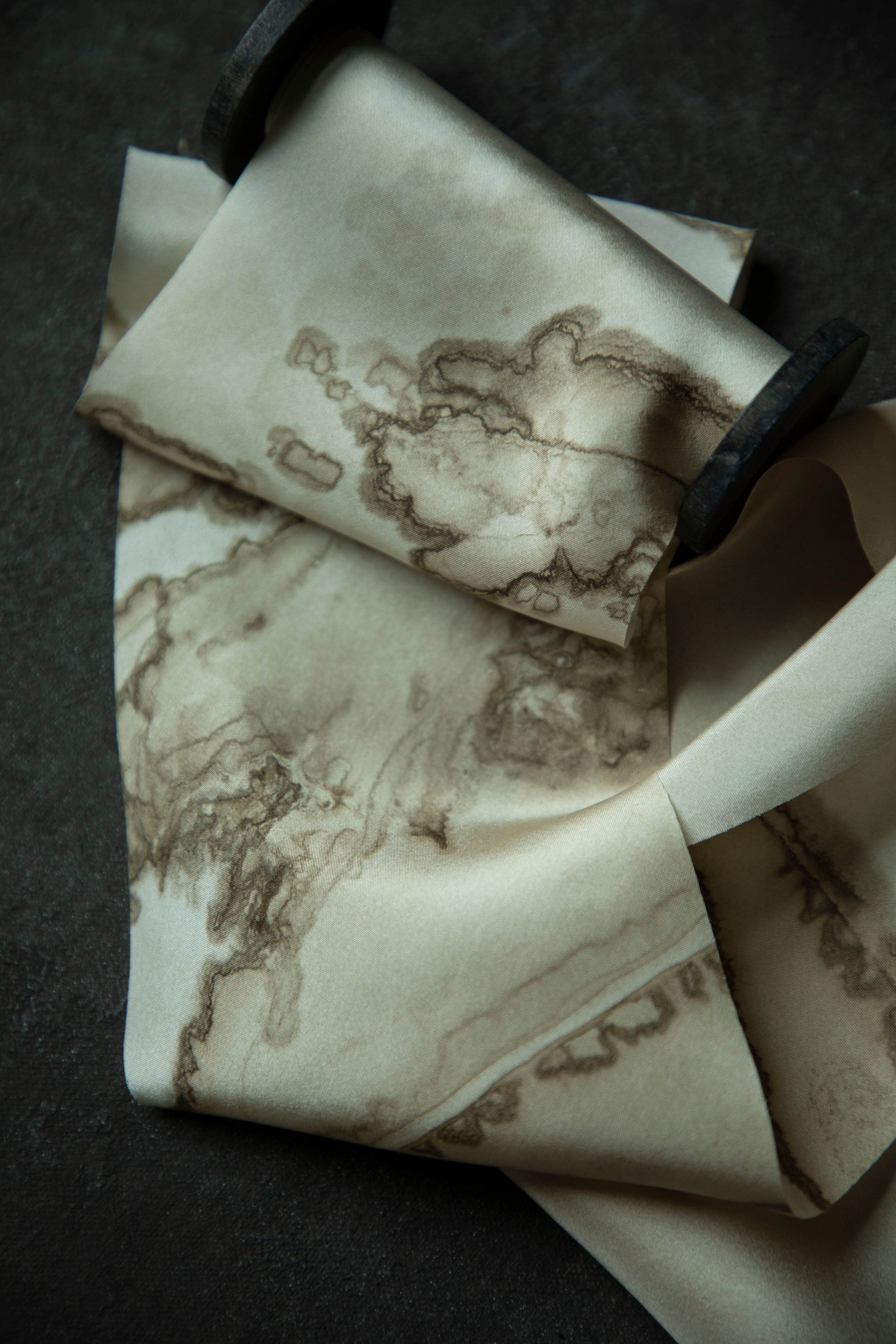 A close-up of a cream-colored silk ribbon with a brown abstract map or ink-blot pattern on a wood spool on a dark textured surface.