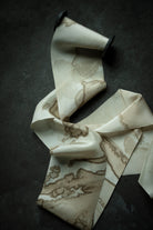 A cream-colored silk ribbon with a brown marble woodgrain pattern, draped elegantly on a dark textured background.