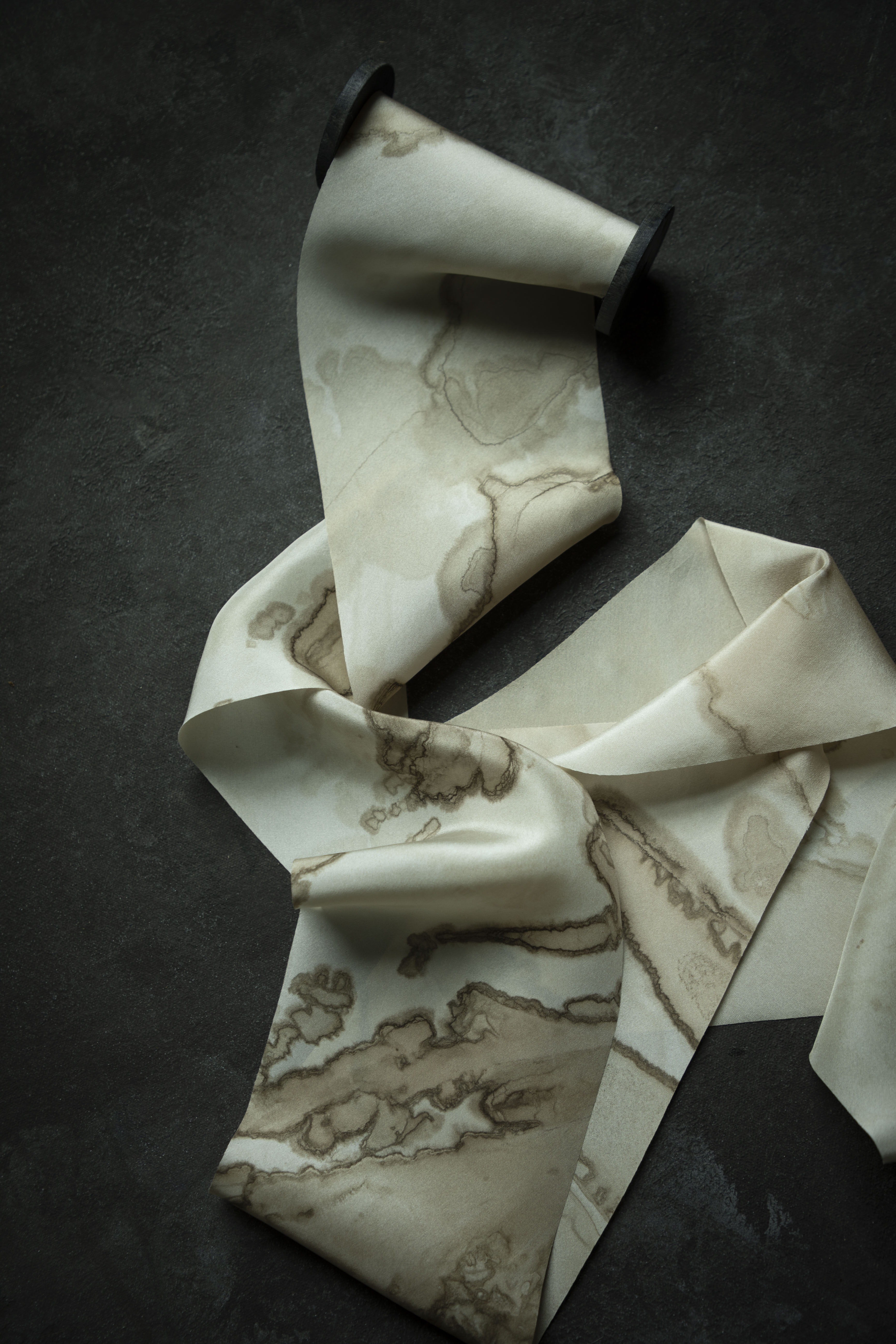 A cream-colored silk ribbon with a brown marble woodgrain pattern, draped elegantly on a dark textured background.