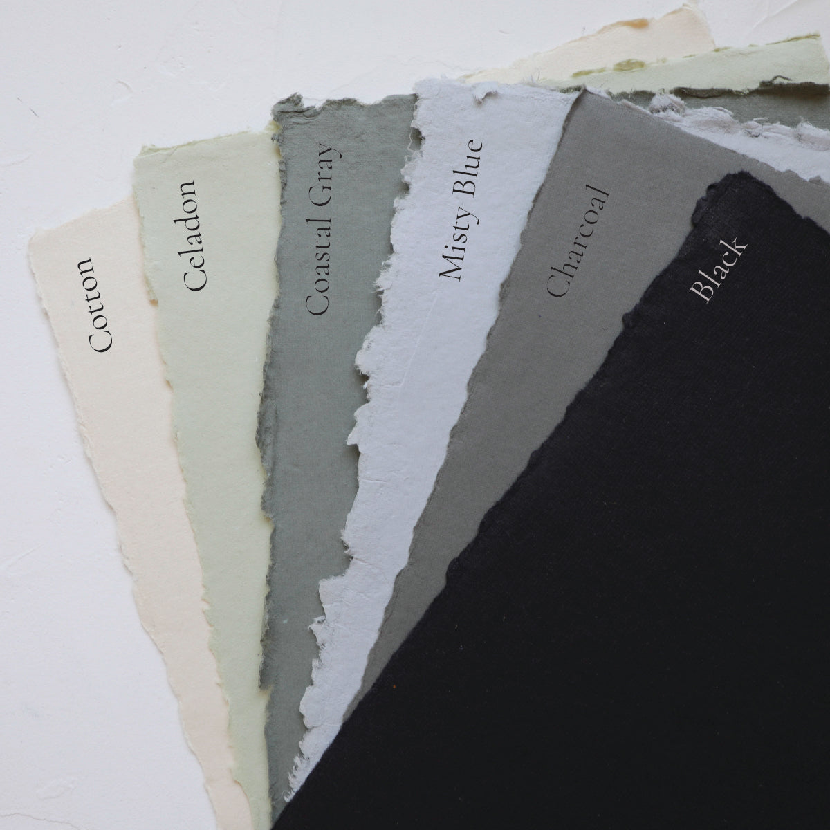 A selection of handmade papers with deckled edges in various shades including white, beige, gray, blue, and black, labeled with their respective color names.